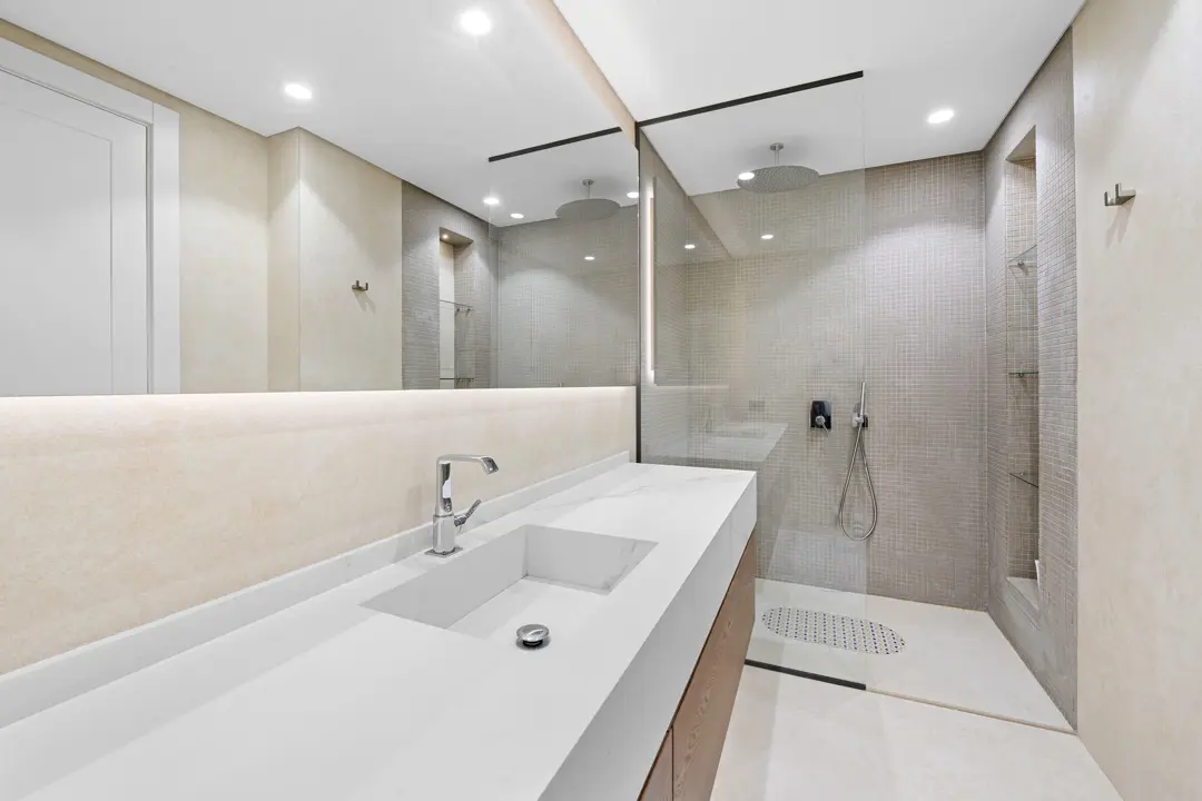 Açık renk tonlarının hakim olduğu, geniş aynalı ve cam duşakabinli modern bir banyo tasarımı. Beyaz tezgah ve ahşap alt dolap kombinasyonu mekana sıcaklık katarken, duş alanındaki mozaik detaylar dikkat çekiyor. Gömme aydınlatmalar sayesinde ferah ve aydınlık bir atmosfer yaratılmış.