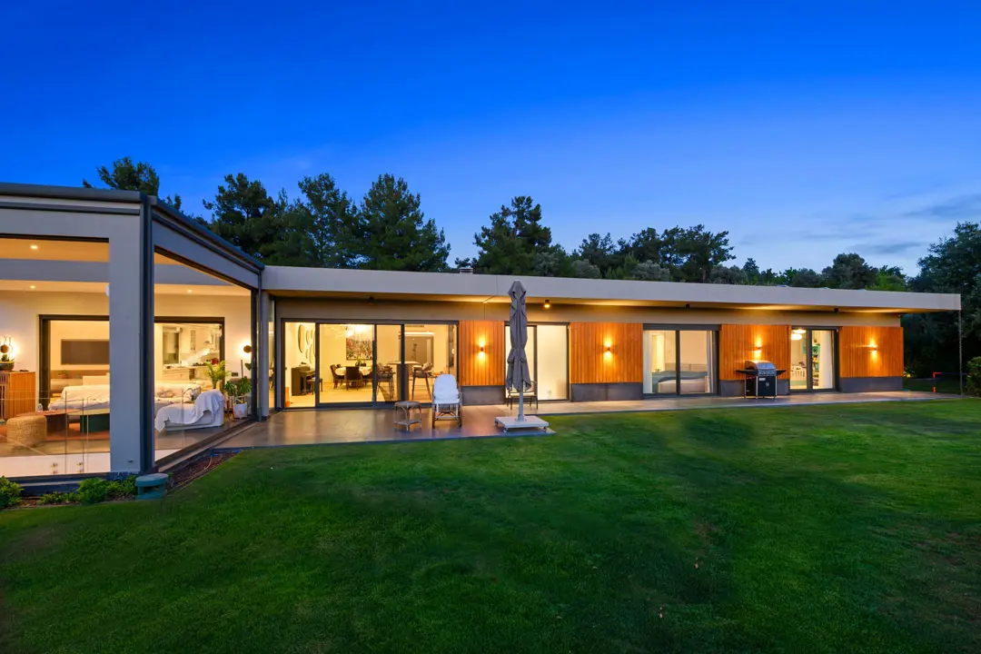 Geniş bir çim bahçeye açılan, tek katlı modern villanın akşam karanlığındaki dış cephe görünümü. Ahşap kaplama detaylar, geniş cam sürgülü kapılar ve sıcak tonlu dış mekan aydınlatmalarıyla davetkar bir atmosfer yaratılmış. İç mekanın sıcak ışığı, dışarıdaki huzurlu doğa manzarasıyla bütünleşiyor.