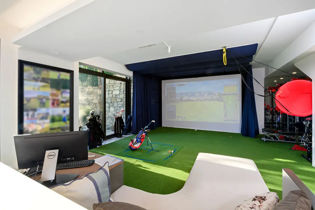 Lüks villanın içinde yer alan kapalı golf simülatörü odası, yeşil suni çim zemin ve büyük bir projeksiyon ekranı ile dikkat çekiyor. Mekanda ayrıca bir çalışma masası, rahat bir oturma alanı ve spor aletleri bulunuyor. Geniş cam pencerelerden sızan doğal ışık, aydınlık ve ferah bir atmosfer yaratıyor.