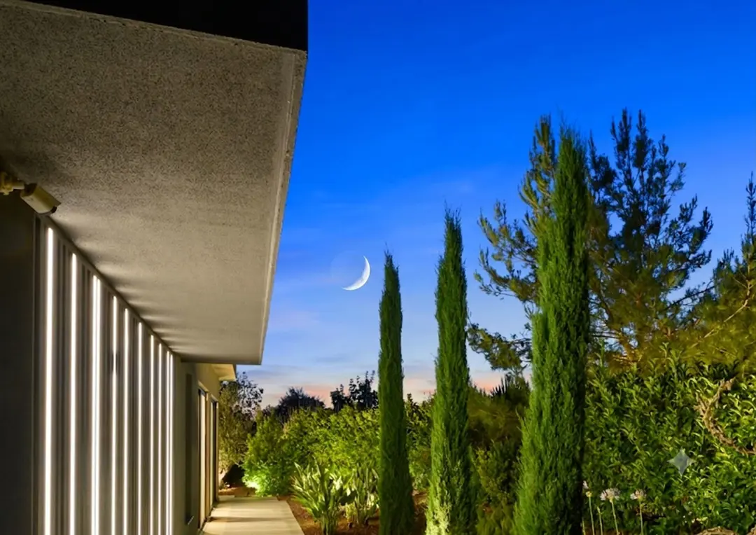 Lüks villanın modern dış cephesi, duvardaki dikey LED aydınlatmaların yarattığı ritmik ışık oyunlarıyla dikkat çekiyor. Geniş saçaklı beton tavanın altındaki yürüyüş yolu, uzun servi ağaçları ve zengin peyzajla bütünleşerek doğayla iç içe bir tasarım sunuyor. Alacakaranlık gökyüzündeki zarif hilal manzarası, mekânın huzurlu ve büyüleyici atmosferini tamamlıyor.