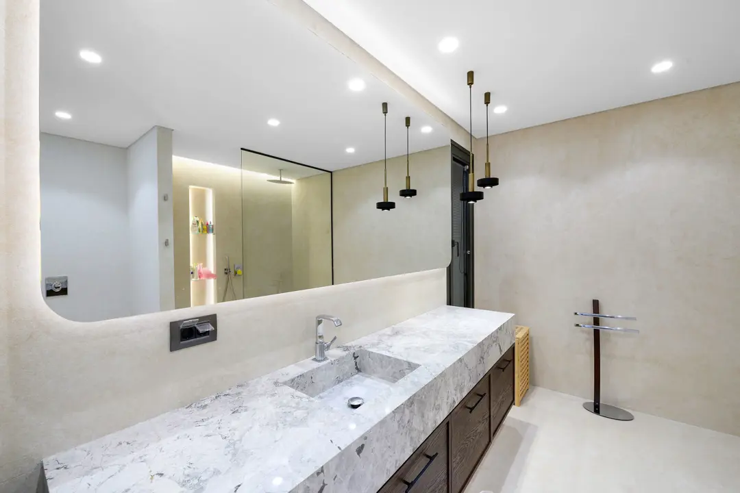 Açık renkli doğal taş duvar kaplamaları ve gri damarlı mermer tezgah ile tasarlanmış modern bir lüks banyo. Geniş, kavisli aynanın arkasından sızan gizli aydınlatma ve sarkıt lambalar mekana sıcak bir atmosfer katıyor. Koyu renkli ahşap dolaplar, mermer ve taşın soğukluğunu dengeleyerek zarif bir kontrast yaratıyor.