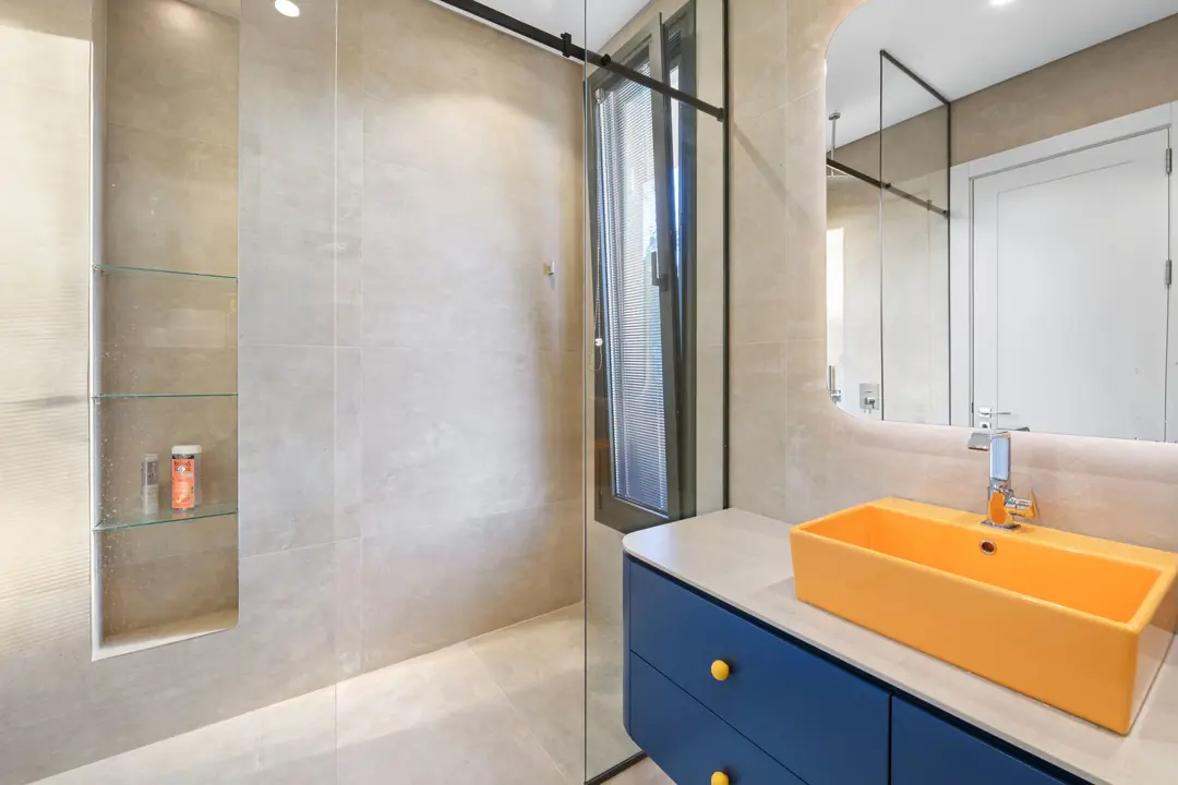 Bu modern banyoda, bej tonlarındaki büyük ebatlı seramik duvar ve zemin kaplamaları, canlı sarı tezgah üstü lavabo ve lacivert banyo dolabı ile çarpıcı bir kontrast oluşturuyor. Siyah profilli cam duşakabin ve niş içindeki cam raflar, mekana şık ve işlevsel bir dokunuş katarken, ayna arkasından gelen gizli aydınlatma ferah bir atmosfer yaratıyor. Pencereden süzülen doğal ışık, renkli detayları daha da vurguluyor.