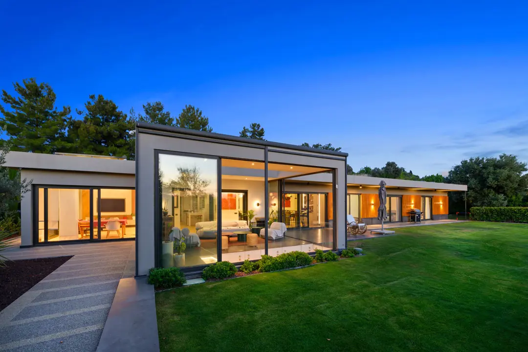 Geniş çim bahçeye açılan, büyük cam cephelere sahip modern tek katlı lüks villa tasarımı. Akşamüstü saatlerinde iç mekandaki sıcak aydınlatmalar, yapının şeffaf ve davetkar atmosferini vurgularken, ahşap detaylar ve koyu renkli doğramalar mimariye derinlik katıyor. İç ve dış mekan arasındaki sınırı kaldıran geniş sürgülü cam kapılar, doğayla bütünleşik bir yaşam alanı sunuyor.