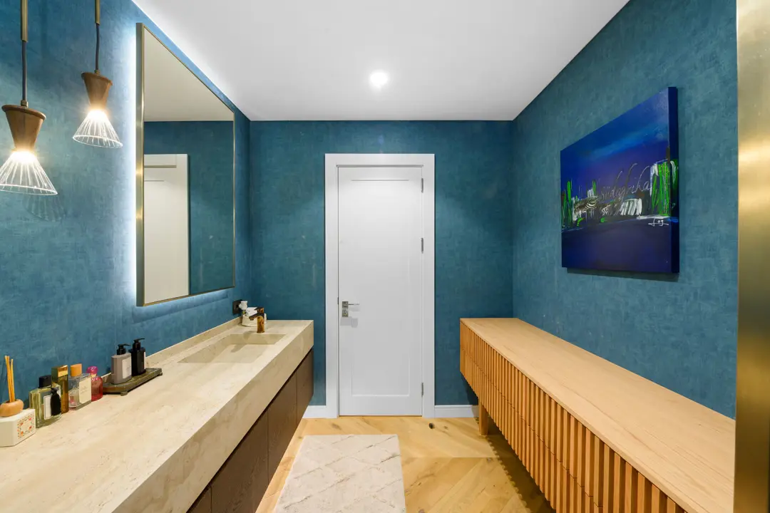 Mavi dokulu duvar kağıtları ve ahşap detayların öne çıktığı modern bir lüks banyo tasarımı. Traverten tezgah, şık sarkıt aydınlatmalar ve duvardaki soyut tablo mekana sanatsal ve sıcak bir atmosfer katıyor. Balıksırtı ahşap zemin ve beyaz kapı ile ferah bir görünüm sağlanmış.