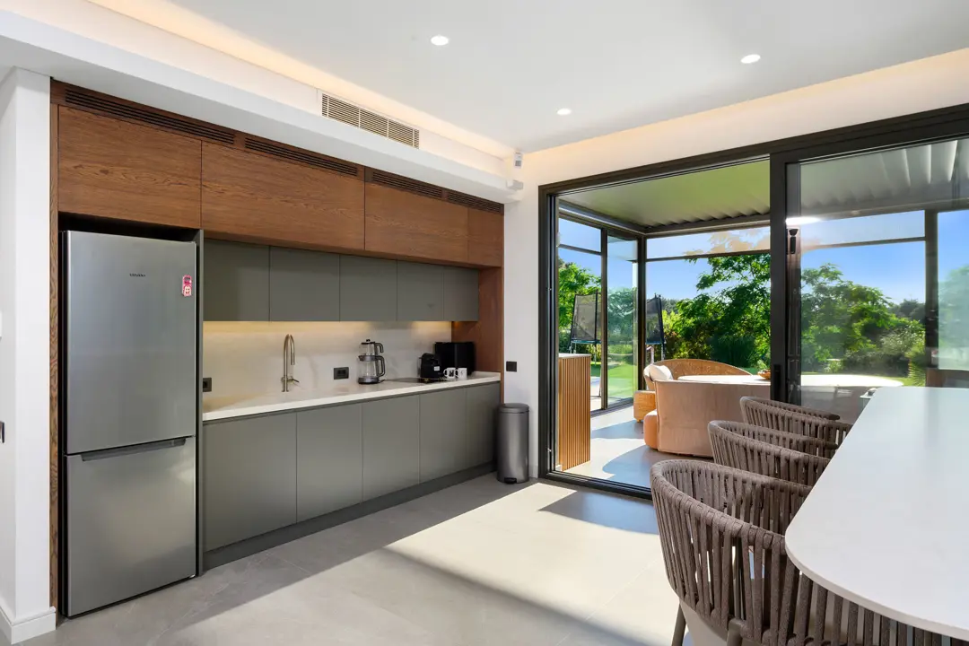 Ahşap ve mat gri dolapların uyum içinde kullanıldığı modern açık plan mutfak. Geniş cam sürgülü kapılar sayesinde doğal ışıkla aydınlanan mekan, bahçe manzarasıyla bütünleşiyor. Hasır sandalyeli ada tezgah, şık ve davetkar bir atmosfer yaratıyor.