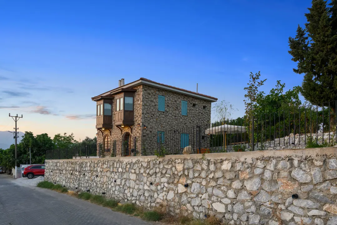 Gün batımı ışığıyla aydınlanan mavi panjurlu ve geleneksel mimarili İzmir Torbalı taş ev projesinin dış mekan fotoğrafı.