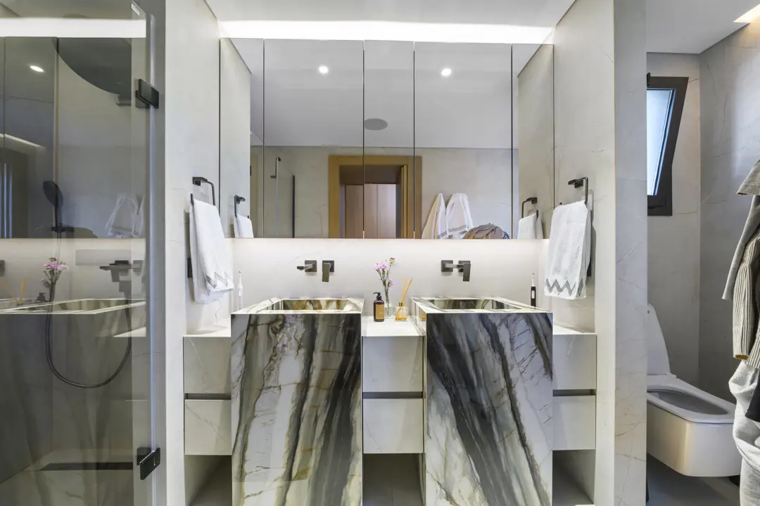 Ebeveyn banyosunda yeşil damarlı mermer çift lavabo, aydınlatmalı ayna dolabı ve cam duşakabin, villa iç mekân.