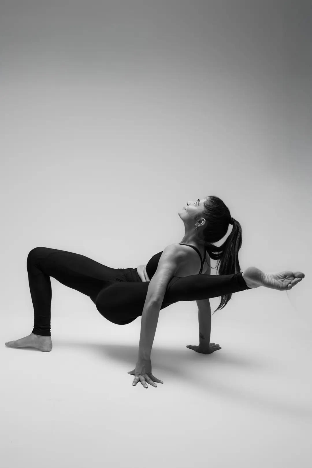 İzmir Menemen'deki stüdyoda açık gri fon önünde zorlu bir asana pozu sergileyen yoga eğitmeni Melis'in siyah beyaz yoga fotoğrafı.